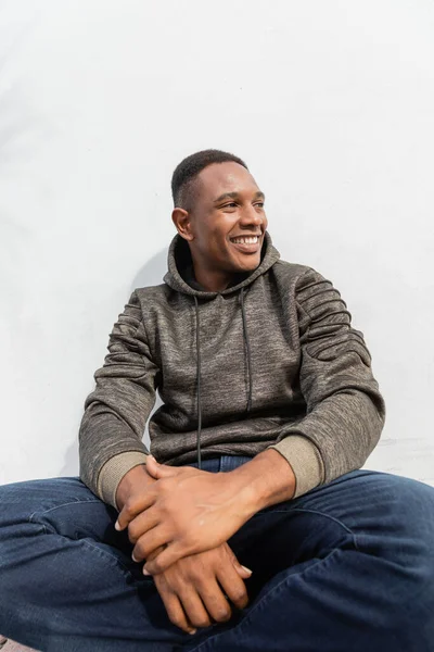 Pleased african american man in jeans and hoodie sitting near white wall — Stock Photo