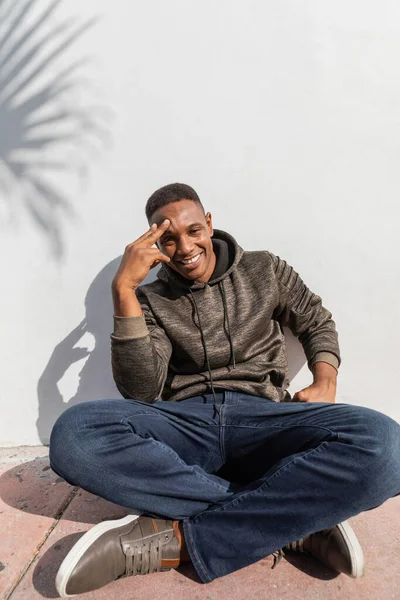 Uomo afroamericano sorridente in jeans e felpa con cappuccio seduto vicino al muro bianco — Foto stock