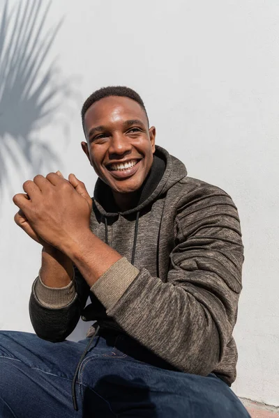 Gioioso uomo africano americano in jeans e felpa con cappuccio sorridente vicino al muro bianco — Foto stock