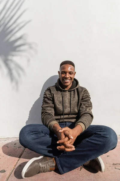 Homme afro-américain insouciant en jeans et sweat à capuche assis près du mur blanc — Photo de stock