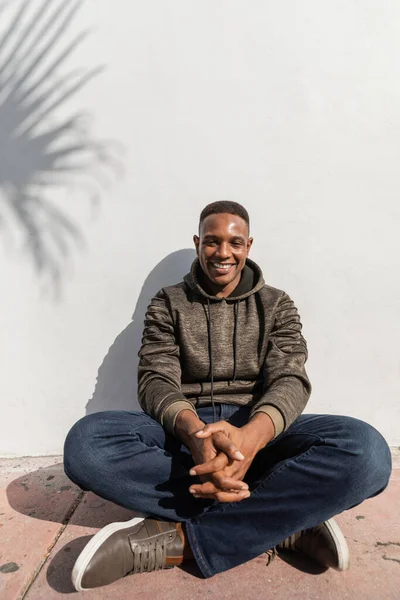 Carefree african american man in jeans and hoodie sitting near white wall — Stock Photo