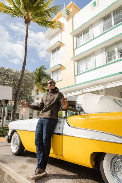 MIAMI, FLORIDE, ÉTATS-UNIS - 15 DÉCEMBRE 2022 : heureux homme afro-américain portant des lunettes de soleil près d'une voiture rétro jaune — Photo de stock