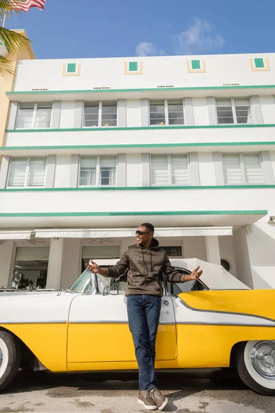 МИАМИ, ФЛОРИДА, США - 15 ДЕКАБРЯ 2022: довольный африканский американец в солнцезащитных очках, стоящий рядом с классическим ретро-автомобилем — стоковое фото