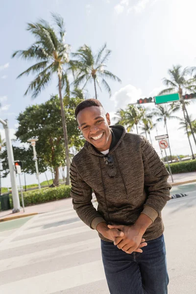 Felice uomo africano americano in felpa con cappuccio sorridente mentre in piedi vicino a palme a Miami — Foto stock