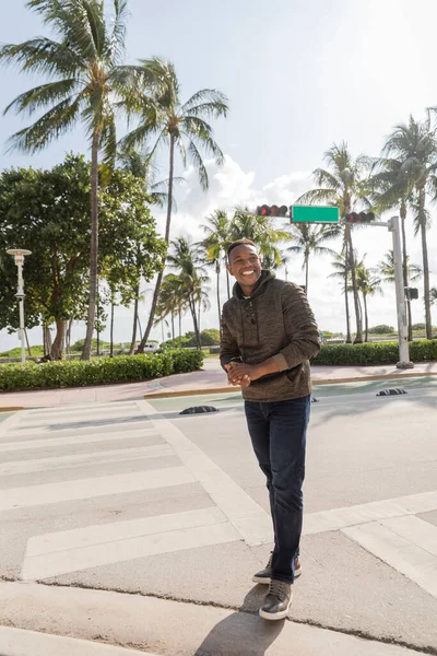 Alegre afroamericano en sudadera con capucha sonriendo mientras está de pie cerca de palmeras en Miami - foto de stock
