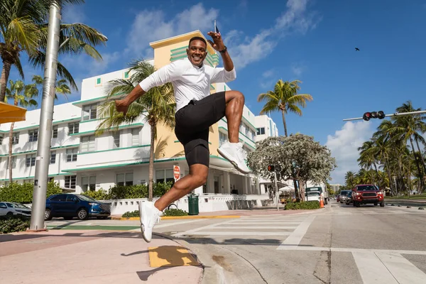 Alegre hombre afroamericano saltando al lado de palmeras y condominio moderno en Miami - foto de stock