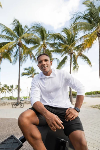 Homme afro-américain joyeux assis sur l'équipement de cross-trainer dans la salle de gym extérieure à Miami — Photo de stock