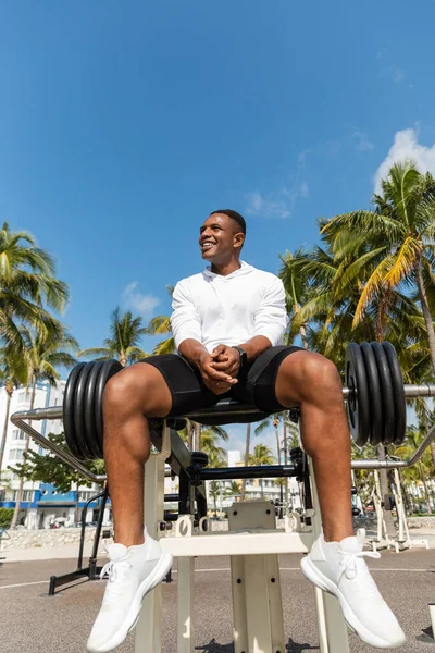 Низкий угол зрения счастливых африканских американских спортсменов, сидящих рядом с барбеллом в открытом тренажерном зале в Майами — стоковое фото