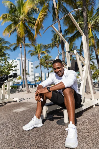 Deportista afroamericano feliz en auriculares inalámbricos descansando después del entrenamiento en la playa de Miami - foto de stock