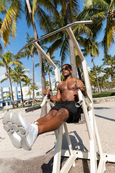 Deportista afroamericano sin camisa haciendo ejercicio en equipo de gimnasio al aire libre en Miami Beach - foto de stock