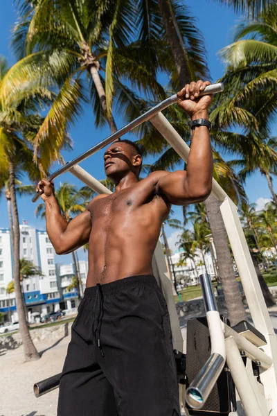 Низкий угол обзора сильного африканского мужчины в шортах, делающего подтягивания на пляже Майами — стоковое фото