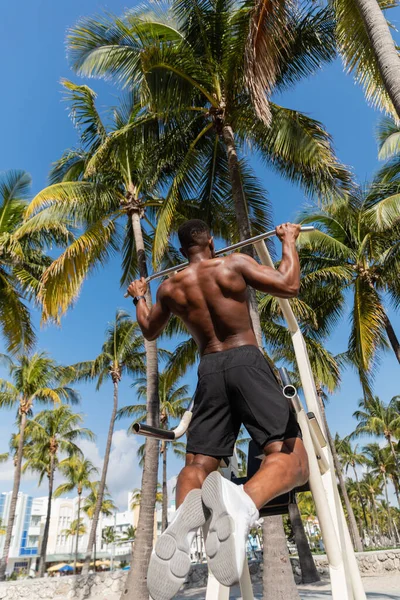 Вид ззаду без сорочки афро-американський спортсмен робить вправи з підтяжками у відкритому тренажерному залі на пляжі Маямі — стокове фото