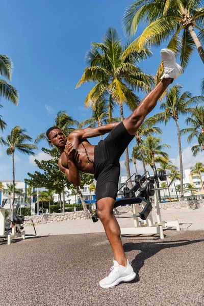 Hombre afroamericano sin camisa en pantalones cortos haciendo ejercicio de patada en Miami Beach - foto de stock