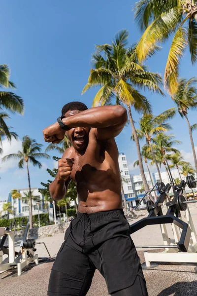 Lutador afro-americano emocional e sem camisa em shorts exercitando na praia de Miami — Fotografia de Stock
