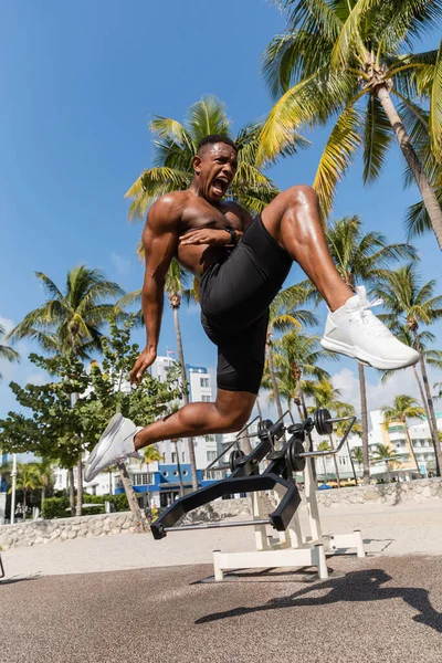 Sportif américain africain émotionnel sautant tout en criant près des palmiers à Miami plage — Photo de stock