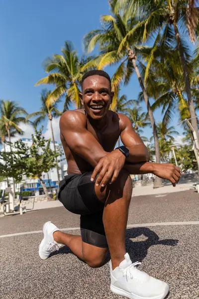 Deportista afroamericano sin camisa sonriendo después de entrenar junto a palmeras en Miami - foto de stock