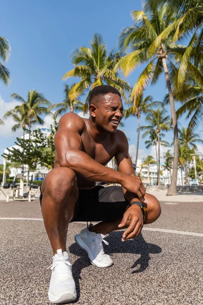 Deportista afroamericano feliz en pantalones cortos y zapatillas de deporte sonriendo después del entrenamiento junto a las palmeras en Miami - foto de stock