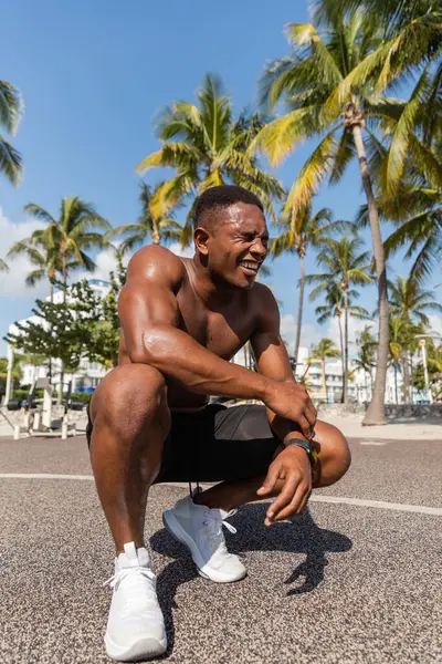 Deportista afroamericano feliz en pantalones cortos y zapatillas de deporte sonriendo después del entrenamiento junto a las palmeras en Miami - foto de stock