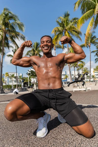 Felice sportivo afroamericano in pantaloncini corti e scarpe da ginnastica che mostra i muscoli dopo l'allenamento sulla spiaggia di Miami — Foto stock