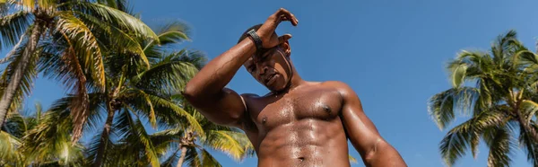 Vue à faible angle de sportif afro-américain en sueur debout à côté de palmiers verts après l'entraînement à Miami, bannière — Photo de stock