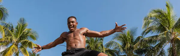 Vista de ángulo bajo del deportista afroamericano emocionado saltando cerca de palmeras verdes en Miami, pancarta - foto de stock