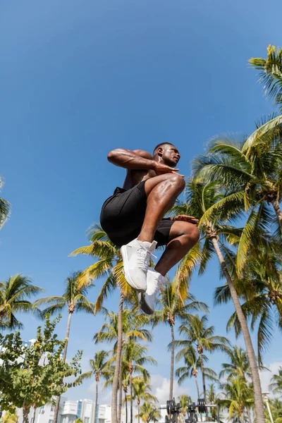 Vue à angle bas d'un sportif afro-américain torse nu en short sautant près de palmiers à Miami — Photo de stock
