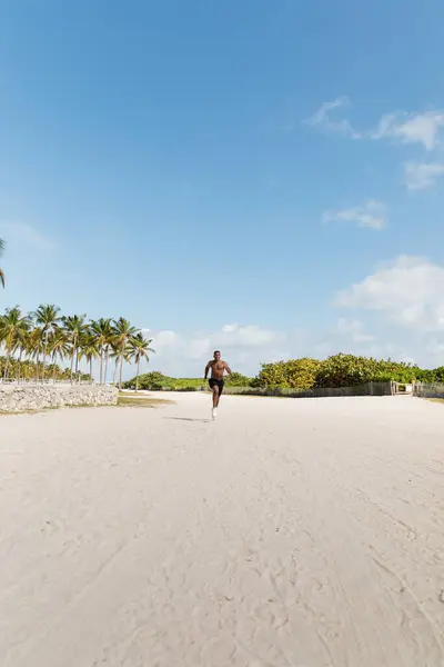 Hombre afroamericano deportivo y sin camisa en pantalones cortos que corren sobre arena en Miami Beach - foto de stock