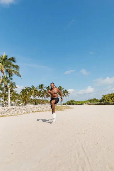 Uomo afroamericano senza maglietta in pantaloncini che corre sulla sabbia accanto alle palme verdi della spiaggia di Miami — Foto stock