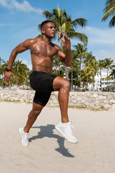 Atlético afroamericano hombre en pantalones cortos y zapatillas de deporte corriendo en Miami Beach - foto de stock