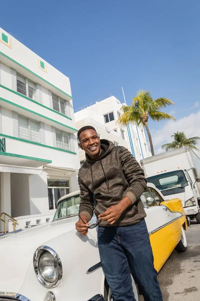 MIAMI, FLORIDE, ÉTATS-UNIS - 15 DÉCEMBRE 2022 : heureux homme afro-américain portant des lunettes de soleil et se tenant près d'une voiture d'époque — Photo de stock