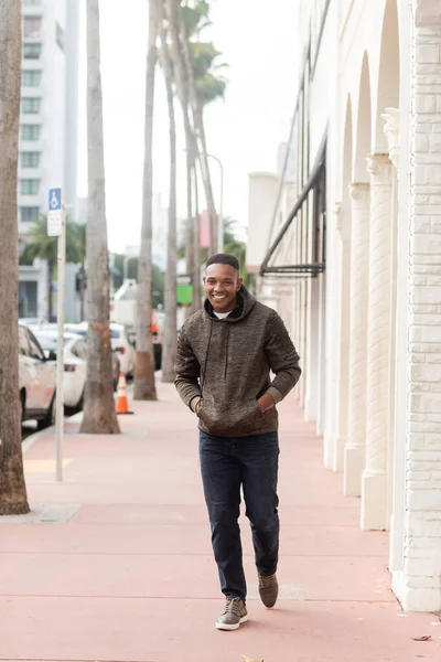 Longitud completa de hombre americano africano alegre en sudadera con capucha y jeans caminando con las manos en los bolsillos en Miami - foto de stock