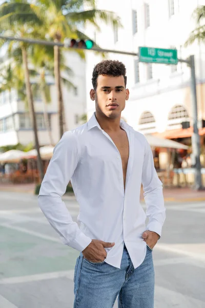Homem cubano de boa aparência em camisa branca e jeans de mãos dadas em bolsos na rua turva em Miami — Fotografia de Stock