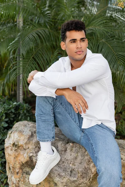 Junger und stylischer kubanischer Mann in weißem Hemd und Jeans schaut weg, während er auf einem Stein in Miami sitzt — Stockfoto