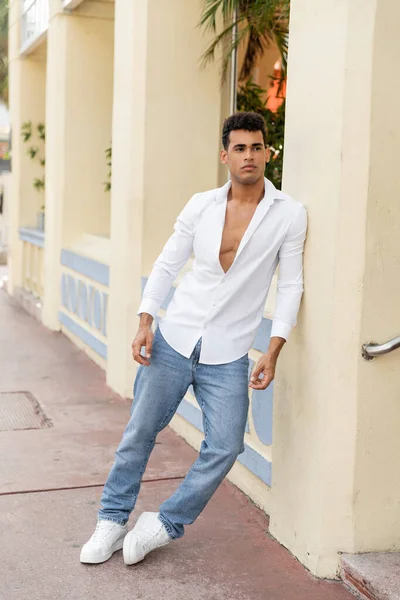 Homem cubano bonito na camisa e jeans olhando afastado perto do edifício na rua urbana em Miami — Fotografia de Stock