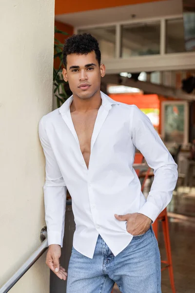 Jeune homme cubain en chemise blanche et jeans posant et regardant la caméra près du bâtiment flou à Miami — Photo de stock