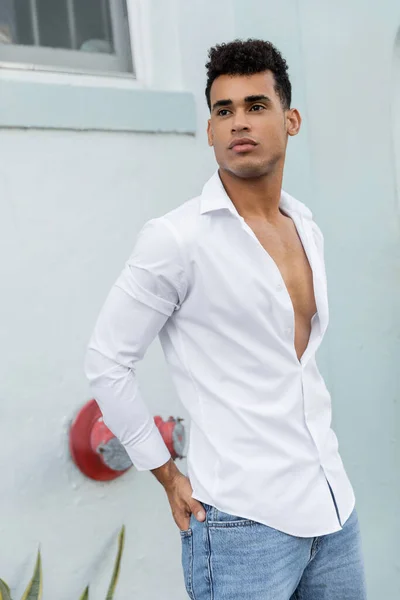 Homem cubano jovem confiante em camisa branca posando enquanto segurava a mão no bolso de jeans em Miami — Fotografia de Stock