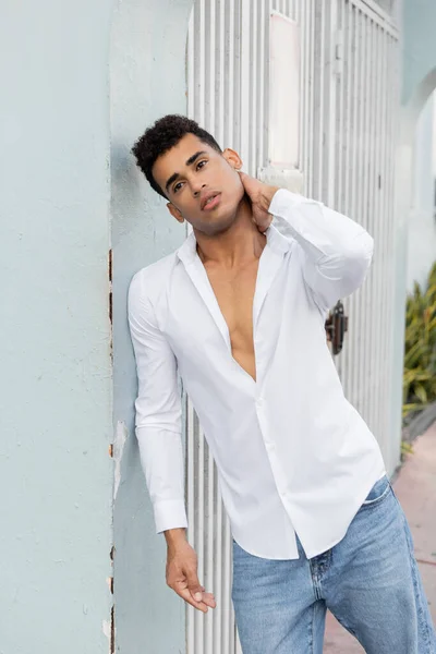 Модный кубинский мужчина в белой рубашке и джинсах трогает шею, стоя возле здания в Майами — стоковое фото