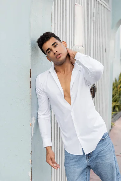 Fashionable cuban man in white shirt and jeans touching neck while standing near building in Miami — Stock Photo