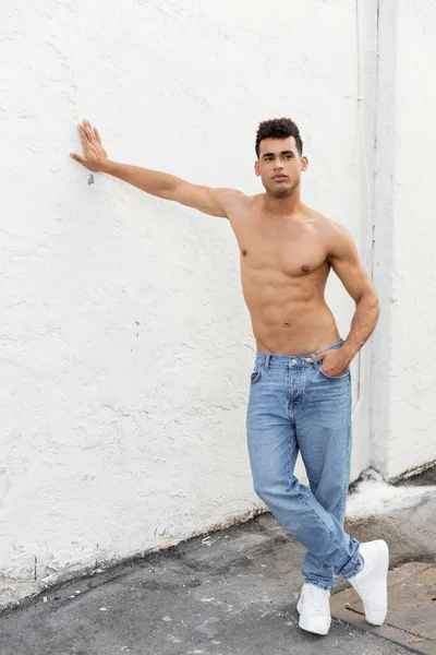Full length of strong and shirtless young cuban man in blue jeans posing and touching wall in Miami — Stock Photo