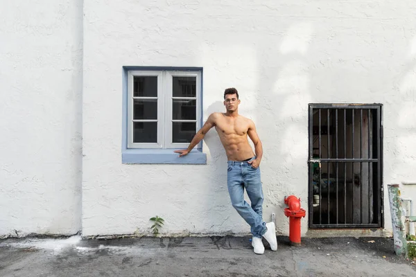 Full length of shirtless young cuban man in jeans and eyeglasses standing near white wall in Miami — Stock Photo