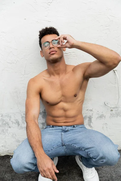 Homem cubano jovem musculoso e sem camisa em jeans azuis tocando óculos de sol redondos — Fotografia de Stock
