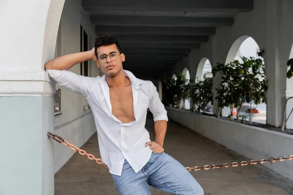 Модний кубинський чоловік у білій сорочці, джинсах та окулярах тримає руку на стегні на міській вулиці в Маямі — стокове фото
