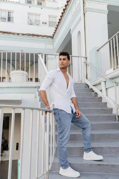 Longitud completa de hombre cubano joven con estilo en camisa blanca y jeans de pie en las escaleras en Miami - foto de stock