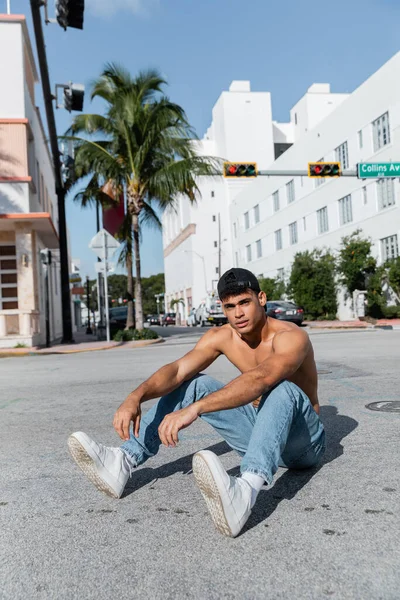 Uomo muscoloso cubano in berretto da baseball e jeans blu seduto su strada a Miami — Foto stock