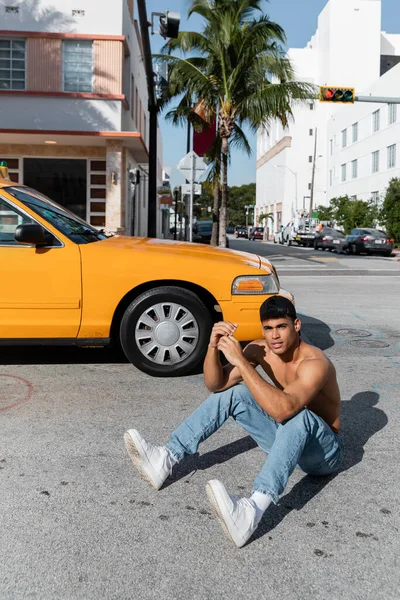 Sexy hombre cubano con cuerpo atlético en gorra de béisbol sentado en la carretera cerca de taxi amarillo en Miami - foto de stock