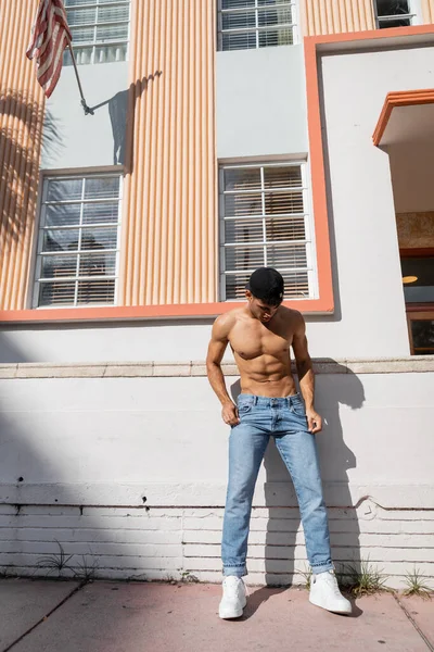 Hombre cubano sin camisa con cuerpo atlético en gorra de béisbol y jeans de pie en la calle en Miami - foto de stock