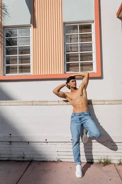 Sexy hombre cubano musculoso con cuerpo atlético en gorra de béisbol y jeans de pie en la calle en Miami - foto de stock