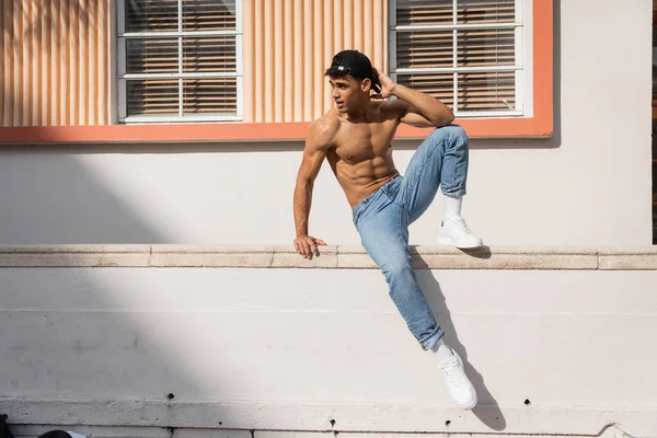 Молодой кубинский мужчина с мускулистым телом позирует в бейсболке и джинсах на улице в Майами летом — стоковое фото