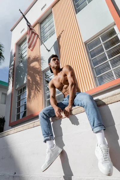 Homme cubain en casquette de baseball, jeans et baskets assis sur le parapet de la construction avec drapeau américain — Photo de stock