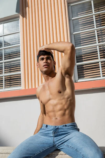 Athlète jeune homme cubain en casquette de baseball et jeans bleus regardant loin tout en étant assis sur parapet — Photo de stock